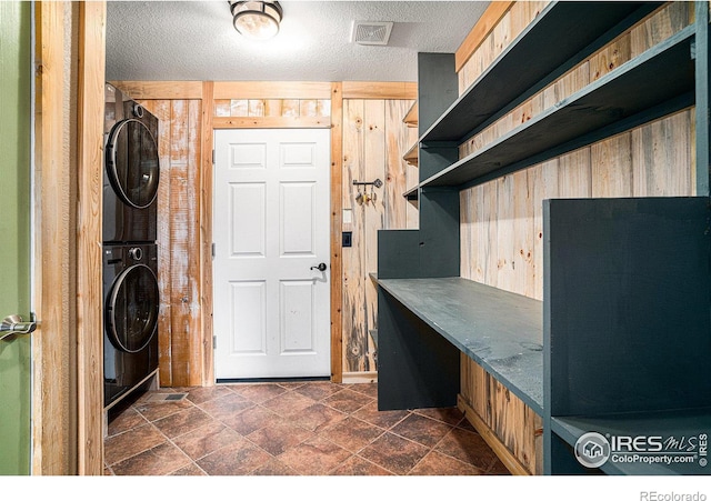 clothes washing area featuring a textured ceiling, stacked washer / drying machine, and wood walls