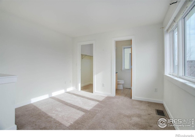 unfurnished bedroom with a walk in closet, ensuite bathroom, multiple windows, and light colored carpet