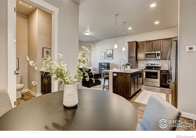 kitchen with dark brown cabinetry, an island with sink, pendant lighting, stainless steel appliances, and decorative backsplash