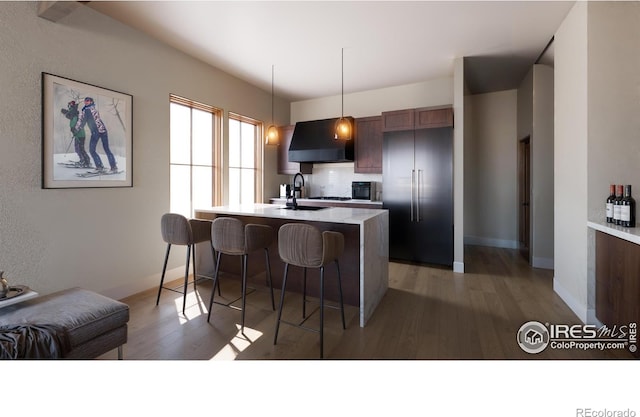 kitchen featuring a center island with sink, wall chimney range hood, hanging light fixtures, decorative backsplash, and a breakfast bar area