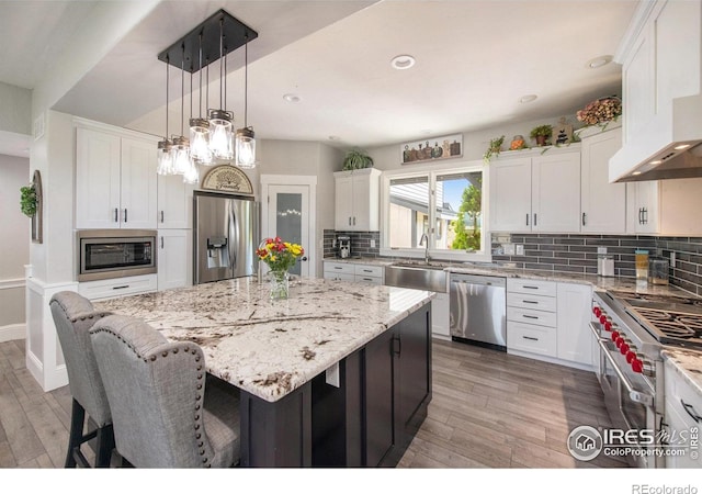 kitchen featuring wall chimney range hood, a kitchen island, pendant lighting, white cabinets, and appliances with stainless steel finishes