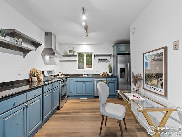 kitchen featuring wall chimney range hood, sink, blue cabinets, light wood-type flooring, and appliances with stainless steel finishes