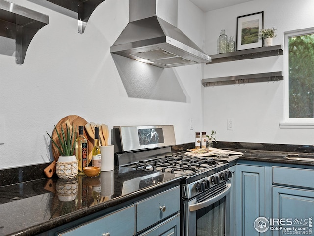 kitchen featuring stainless steel range with gas cooktop, blue cabinets, dark stone countertops, and wall chimney range hood