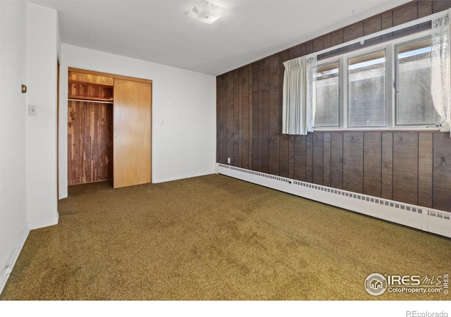 unfurnished bedroom featuring dark colored carpet, baseboard heating, a closet, and wood walls