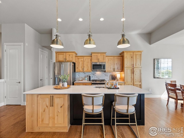 kitchen with light brown cabinets, backsplash, a center island with sink, light wood-type flooring, and appliances with stainless steel finishes