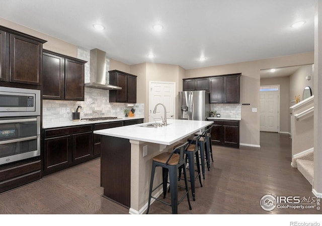 kitchen with sink, wall chimney exhaust hood, a kitchen breakfast bar, an island with sink, and appliances with stainless steel finishes