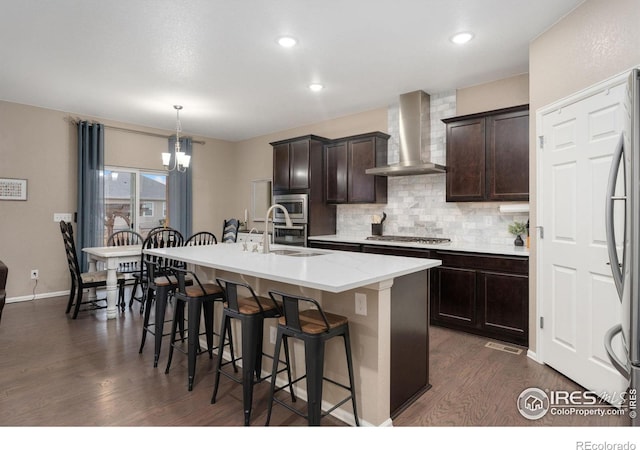 kitchen with an inviting chandelier, a center island with sink, wall chimney exhaust hood, appliances with stainless steel finishes, and decorative light fixtures