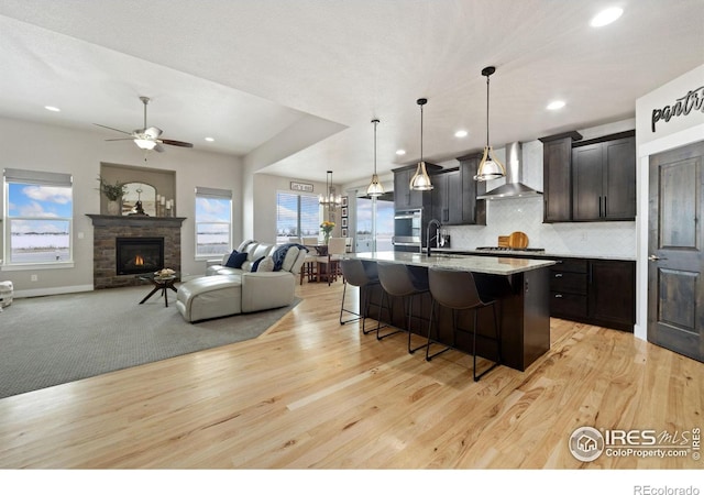 kitchen featuring wall chimney range hood, a kitchen breakfast bar, light hardwood / wood-style flooring, pendant lighting, and a center island with sink