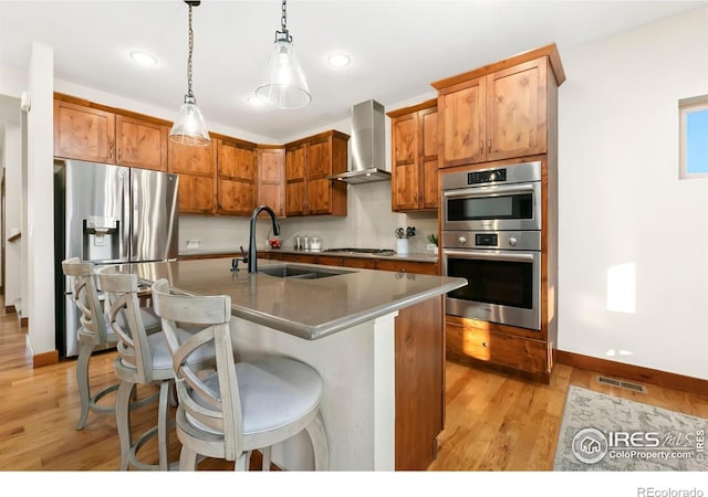 kitchen with an island with sink, stainless steel appliances, decorative light fixtures, sink, and wall chimney exhaust hood