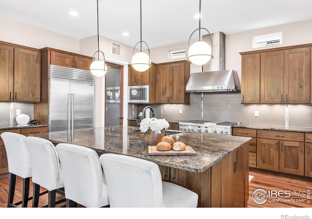 kitchen featuring a kitchen island with sink, wall chimney exhaust hood, decorative light fixtures, and appliances with stainless steel finishes