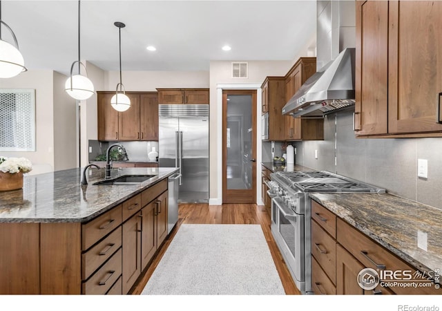 kitchen featuring high end appliances, sink, wall chimney range hood, pendant lighting, and dark stone countertops