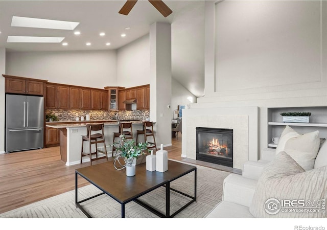 living room with a skylight, ceiling fan, high vaulted ceiling, light hardwood / wood-style floors, and a tiled fireplace