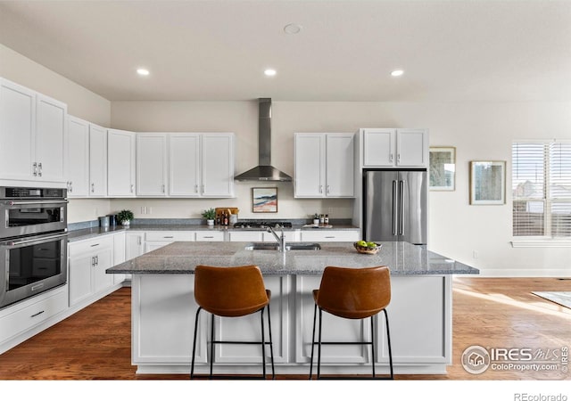 kitchen with white cabinetry, wall chimney range hood, a kitchen island with sink, and appliances with stainless steel finishes