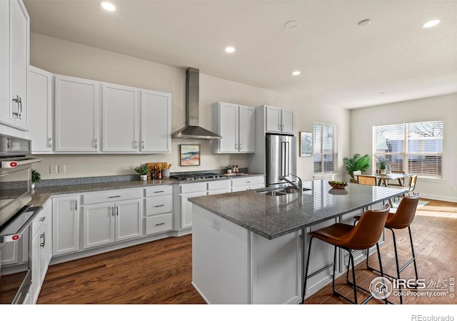 kitchen featuring white cabinets, wall chimney range hood, sink, an island with sink, and stainless steel appliances