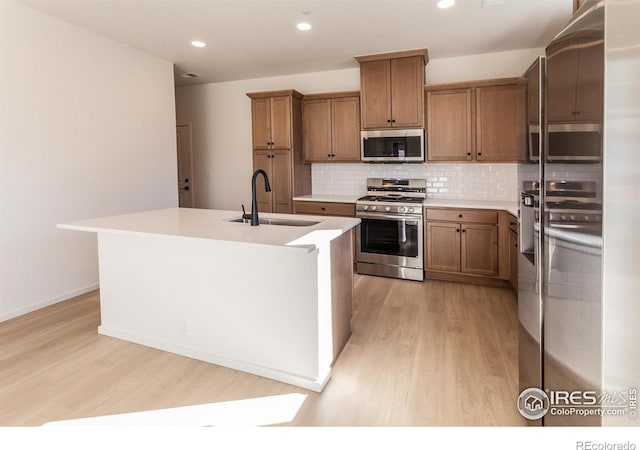 kitchen featuring sink, light hardwood / wood-style flooring, an island with sink, appliances with stainless steel finishes, and tasteful backsplash