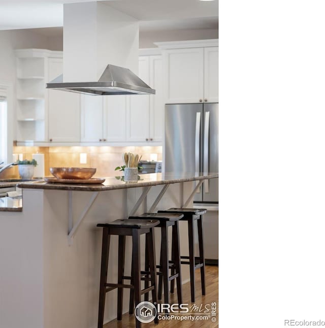 kitchen featuring wall chimney range hood, dark hardwood / wood-style floors, white cabinetry, a breakfast bar area, and stainless steel refrigerator