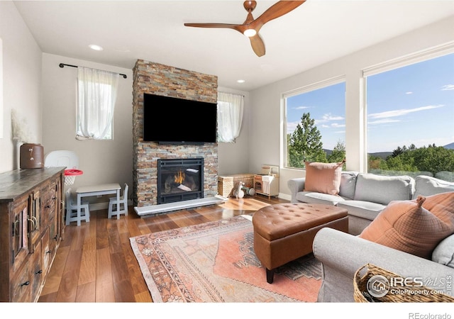 living room with a stone fireplace, ceiling fan, and dark wood-type flooring