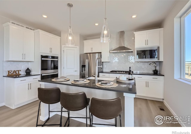 kitchen featuring appliances with stainless steel finishes, tasteful backsplash, a kitchen island with sink, wall chimney range hood, and white cabinets