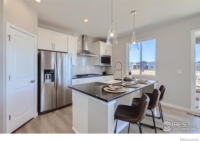 kitchen featuring wall chimney exhaust hood, stainless steel appliances, decorative backsplash, a kitchen island with sink, and white cabinets