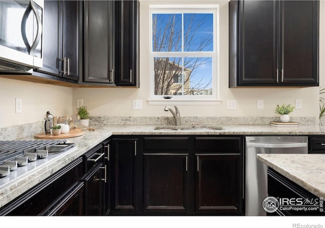 kitchen featuring stainless steel appliances, dark cabinetry, a sink, and light stone countertops