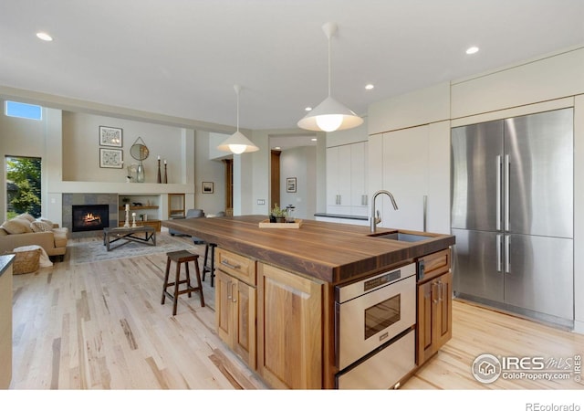 kitchen with a tile fireplace, hanging light fixtures, wooden counters, stainless steel built in refrigerator, and an island with sink