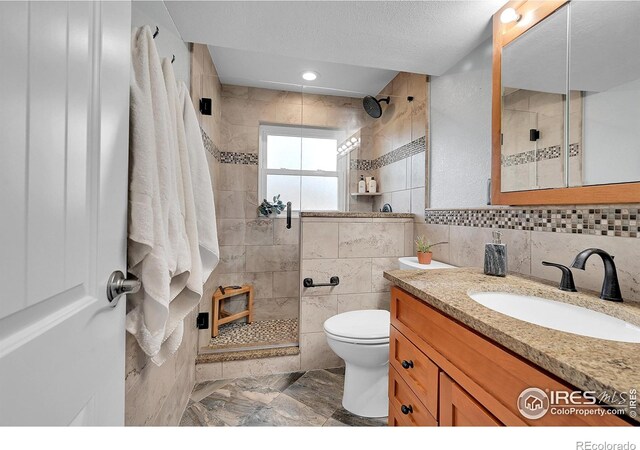 bathroom featuring a textured ceiling, toilet, tiled shower, vanity, and tile walls