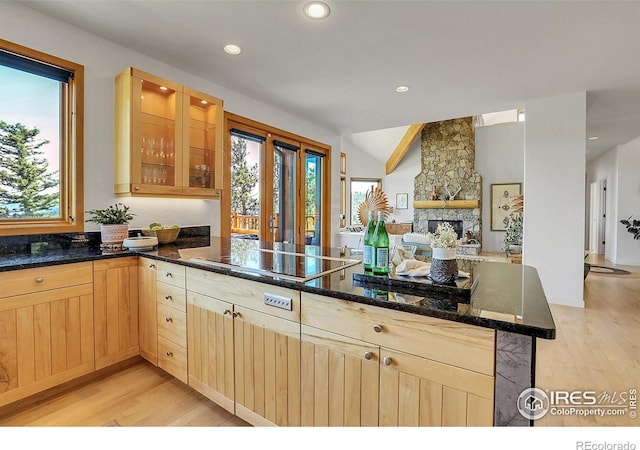 kitchen with light wood-type flooring, kitchen peninsula, a stone fireplace, and dark stone counters