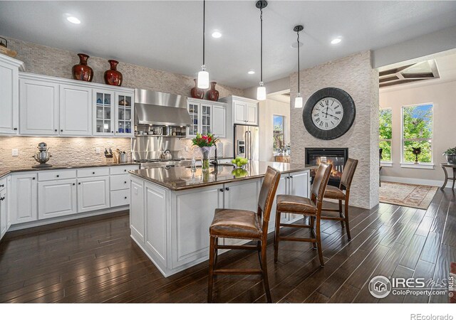 kitchen with white cabinets, hanging light fixtures, wall chimney exhaust hood, an island with sink, and high quality fridge