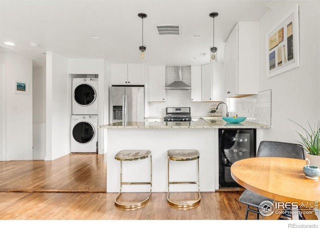 kitchen featuring wall chimney exhaust hood, hanging light fixtures, stainless steel fridge, stacked washer / drying machine, and white cabinets