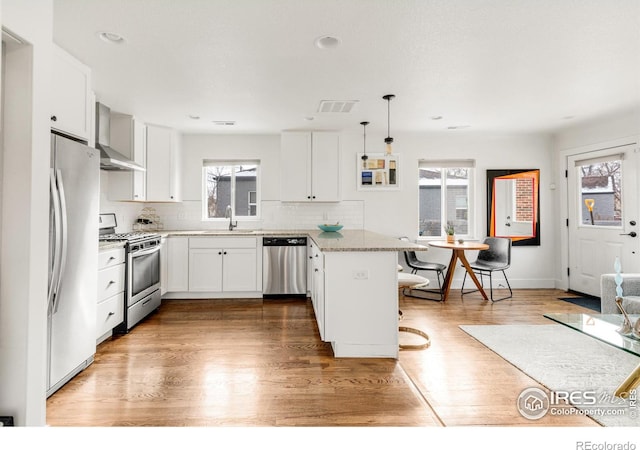 kitchen featuring appliances with stainless steel finishes, wall chimney exhaust hood, sink, decorative light fixtures, and white cabinets