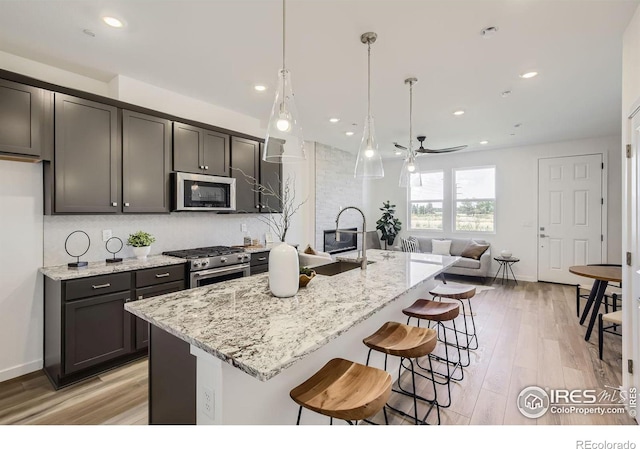 kitchen with stainless steel appliances, decorative light fixtures, ceiling fan, light stone countertops, and a kitchen island with sink