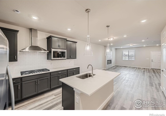 kitchen featuring sink, stainless steel appliances, wall chimney exhaust hood, light hardwood / wood-style flooring, and an island with sink