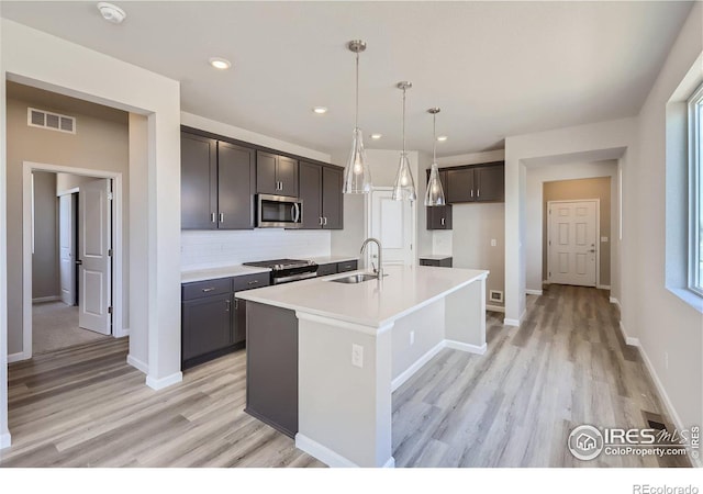 kitchen with appliances with stainless steel finishes, pendant lighting, an island with sink, sink, and light hardwood / wood-style floors