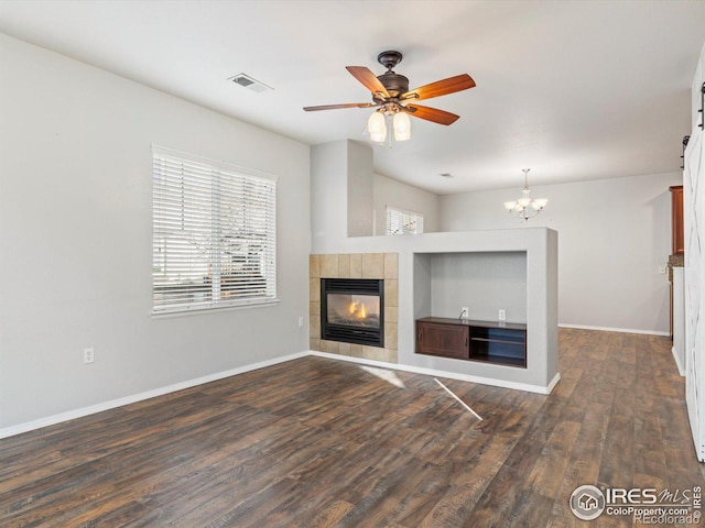 unfurnished living room with ceiling fan with notable chandelier, a tiled fireplace, and dark hardwood / wood-style floors