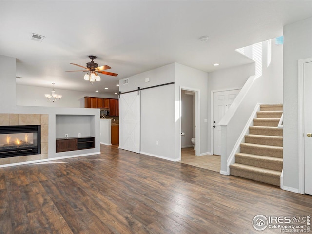 unfurnished living room with ceiling fan with notable chandelier, a tiled fireplace, a barn door, and hardwood / wood-style floors