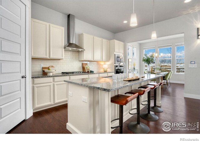 kitchen with white cabinetry, a center island with sink, light stone counters, appliances with stainless steel finishes, and wall chimney range hood