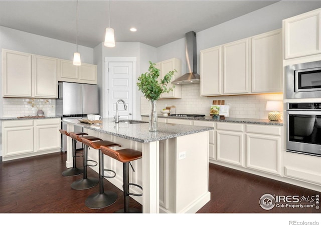 kitchen with a center island with sink, stainless steel appliances, light stone counters, wall chimney range hood, and sink