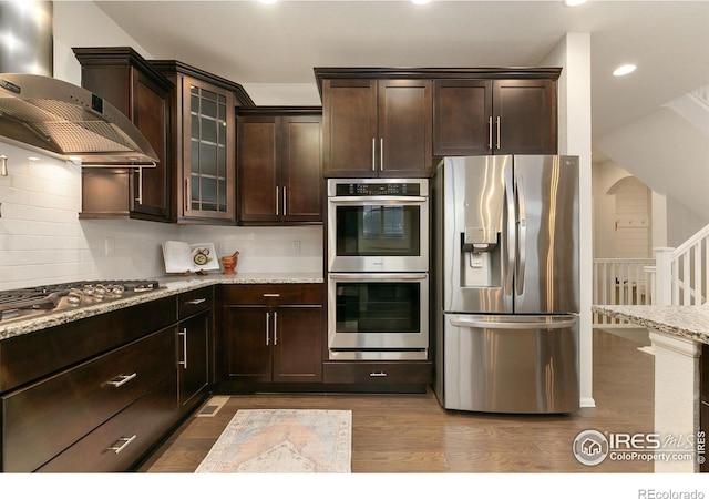 kitchen featuring light stone counters, wall chimney exhaust hood, tasteful backsplash, dark hardwood / wood-style flooring, and appliances with stainless steel finishes