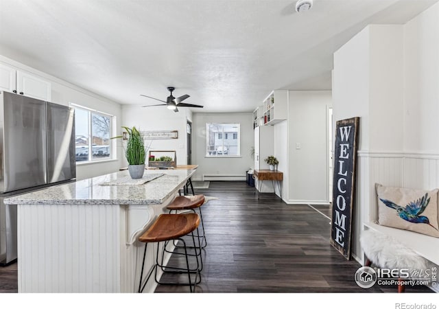 kitchen featuring light stone counters, stainless steel fridge, baseboard heating, a breakfast bar area, and white cabinets