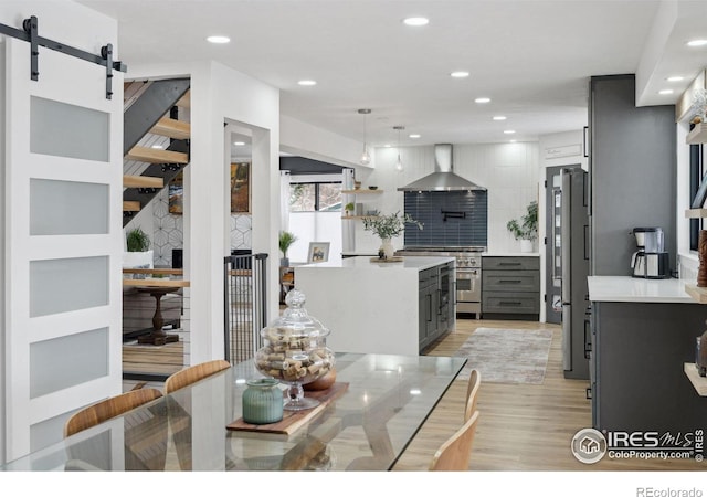 kitchen featuring light countertops, gray cabinetry, a barn door, appliances with stainless steel finishes, and wall chimney exhaust hood