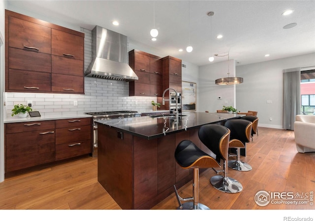 kitchen featuring sink, light hardwood / wood-style floors, an island with sink, hanging light fixtures, and wall chimney range hood