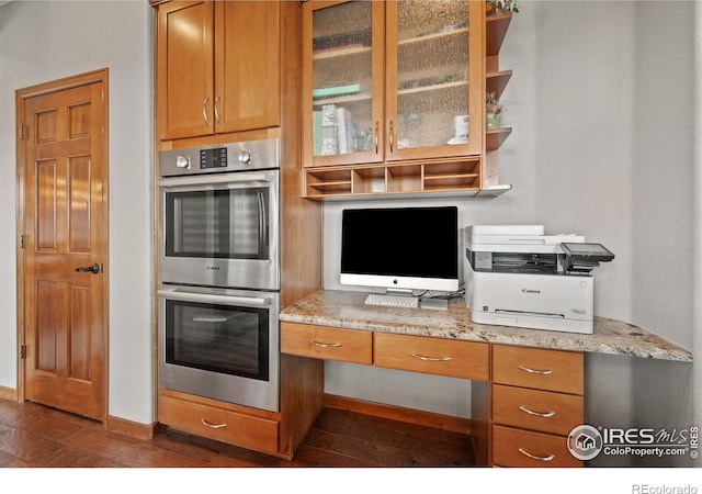 kitchen with light stone counters, dark wood-type flooring, stainless steel double oven, and built in desk