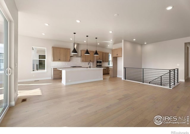 kitchen with wall chimney exhaust hood, light hardwood / wood-style floors, sink, hanging light fixtures, and a center island with sink