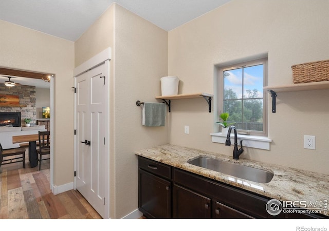 kitchen with a stone fireplace, sink, light stone counters, dark brown cabinets, and light wood-type flooring