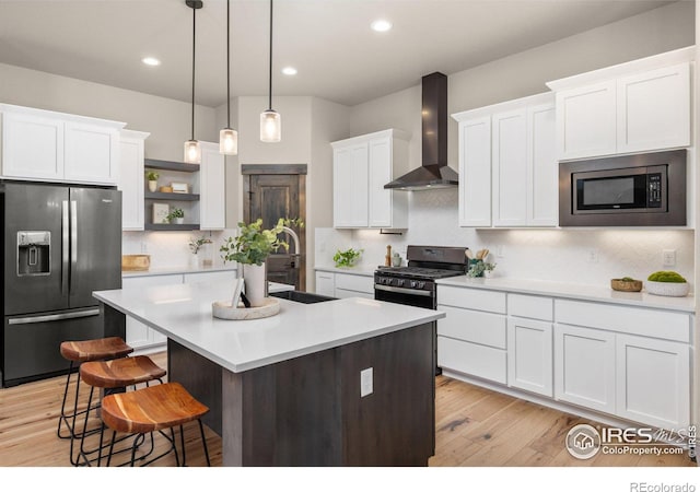 kitchen featuring stainless steel appliances, a kitchen island with sink, hanging light fixtures, wall chimney range hood, and white cabinets