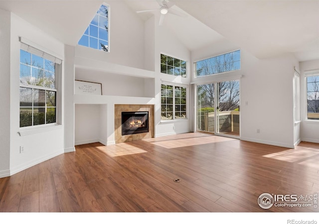 unfurnished living room featuring ceiling fan, a tiled fireplace, wood finished floors, high vaulted ceiling, and baseboards