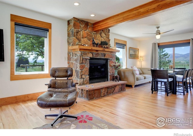 living room featuring ceiling fan, wood-type flooring, beamed ceiling, and a stone fireplace