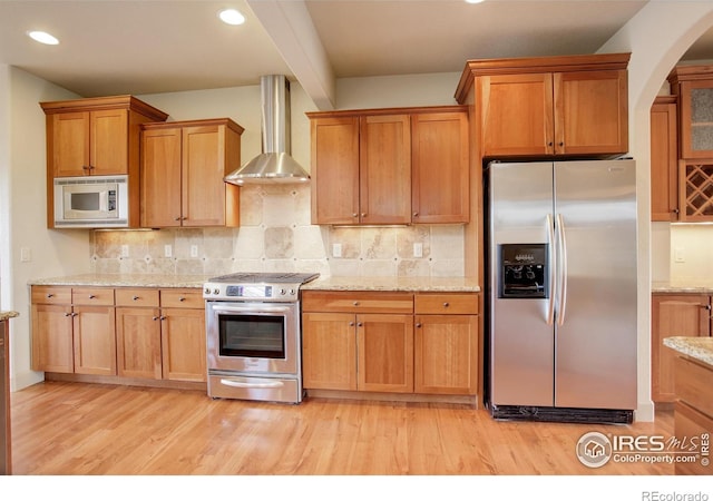 kitchen featuring tasteful backsplash, light wood-type flooring, stainless steel appliances, wall chimney exhaust hood, and light stone counters