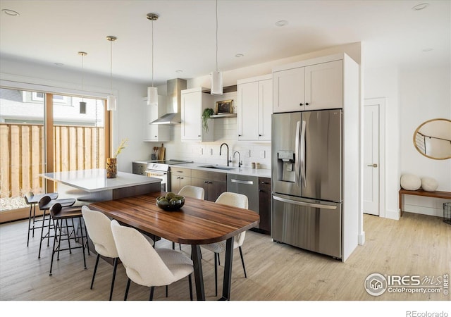 kitchen with white cabinetry, stainless steel appliances, hanging light fixtures, wall chimney range hood, and sink