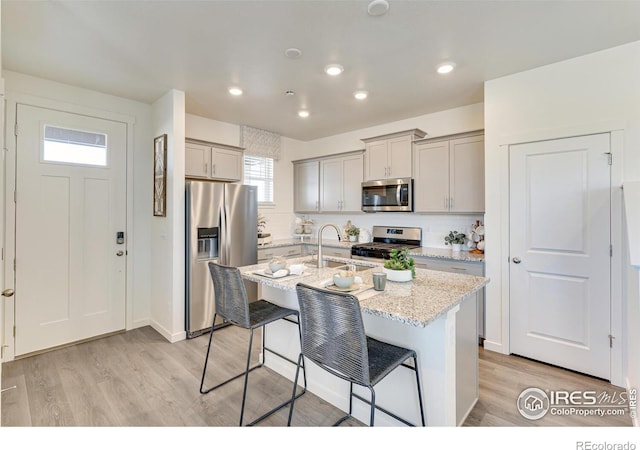 kitchen with a kitchen bar, stainless steel appliances, an island with sink, light stone counters, and light hardwood / wood-style flooring